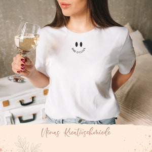 Peut inclure: T-shirt blanc avec un motif de smiley noir qui dit "Wine O'Clock". Une personne tient un verre de vin blanc.