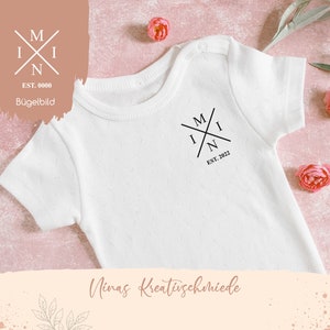 Könnte beinhalten: Ein weißer Baby-Strampler mit einer schwarzen Grafik eines stilisierten Buchstabens 'M', der sich mit den Buchstaben 'I' und 'N' zu einem Kreuz kreuzt. Der Text 'EST. 2022' befindet sich unterhalb der Grafik. Der Strampler liegt auf einem rosa Hintergrund mit Blumen.