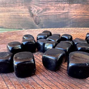 May include: A collection of polished black obsidian stones, each with a smooth, rounded shape. The stones are displayed on a wooden surface, showcasing their glossy, reflective surfaces. The dark color of the stones contrasts with the warm tones of the wood.