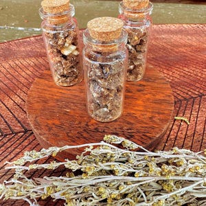 May include: Three small glass bottles with cork stoppers, filled with dried herbs, sit on a wooden surface. A bundle of dried, light green herbs lies in front of the bottles. The background is a mix of red and brown.