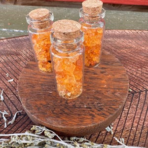 May include: Three small glass bottles filled with orange-colored crystals, each sealed with a cork stopper. The bottles are arranged on a round, wooden surface. Dried herbs are scattered around the base.