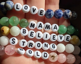 May include: A collection of beaded bracelets with white letter beads spelling out "LOVE", "MAMA", "BELIEVE", "STRONG", and "BOLD". The bracelets feature a variety of colorful gemstone beads, including blue, green, pink, and white.