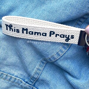 Può includere: Portachiavi in tela bianca con testo nero che recita "This Mama Prays" e l'account social "@happybutmessy".