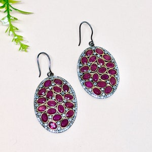 May include: A pair of silver earrings with a cluster of oval-shaped red gemstones surrounded by smaller white gemstones. The earrings have a simple hook closure.