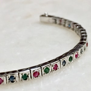 Könnte beinhalten: Ein silbernes Armband mit einer Reihe von bunten Edelsteinen. Die Edelsteine sind in quadratischen Fassungen gefasst und in einem sich wiederholenden Muster aus Rot, Grün und Blau angeordnet.