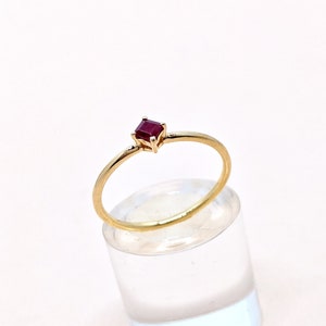 May include: A simple gold band ring with a small, square-shaped ruby gemstone.