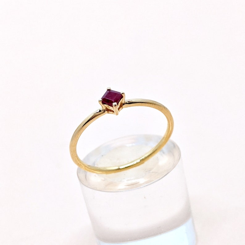 May include: A simple gold band ring with a small, square-shaped ruby gemstone.