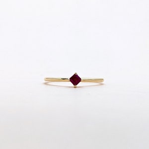 May include: A simple gold band ring with a small square-shaped ruby gemstone in the center.