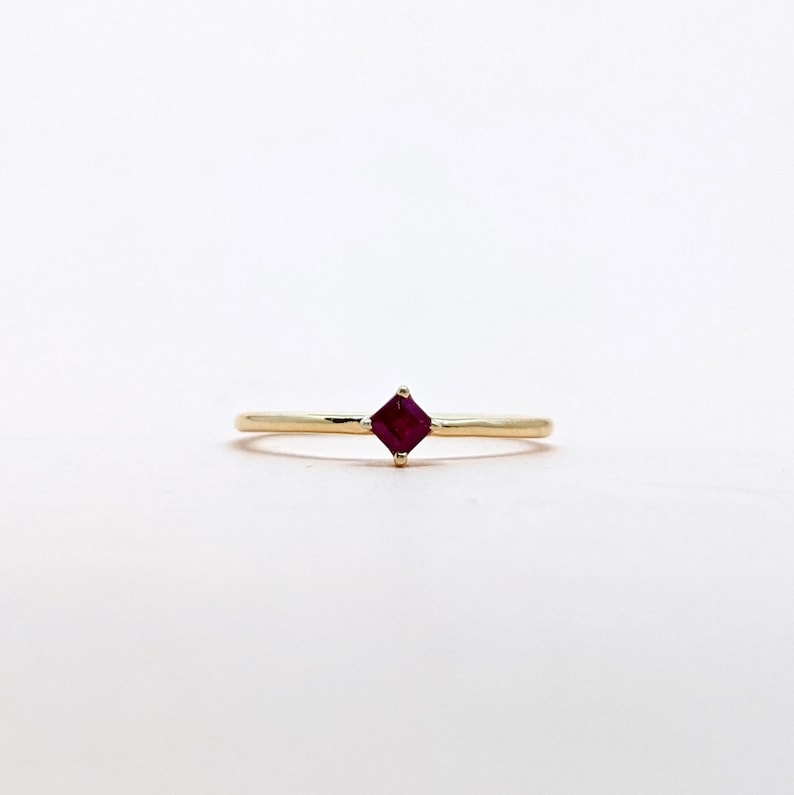 May include: A simple gold band ring with a small square-shaped ruby gemstone in the center.