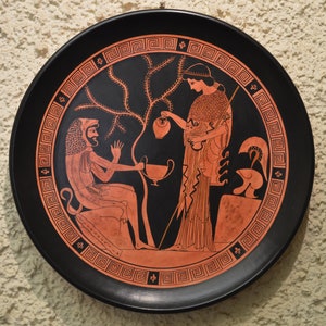 May include: A black ceramic plate with a red and white design depicting a scene from Greek mythology. A woman pours liquid from a jug into a cup held by a man with a lion's head.