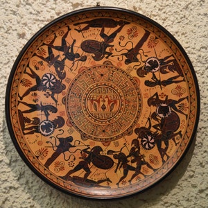 May include: A round ceramic plate with a black rim and a brown background. The plate is decorated with a black and white design of Greek warriors fighting. The design includes a central circle with a geometric pattern and the words "ENKAI" and "KORNEIA".