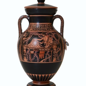 May include: A black and orange Greek amphora with a lid. The amphora is decorated with a scene of a reclining figure and a bird.