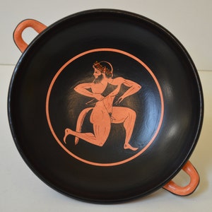Puede incluir: Un cuenco de cerámica negra con un diseño rojo-naranja de una figura con una corona en la cabeza. La figura está representada en una pose de carrera.