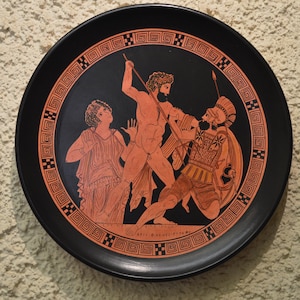 Könnte beinhalten: Ein schwarzer Keramikteller mit einem roten und weißen Design, das eine Szene aus der griechischen Mythologie darstellt. Die Szene zeigt drei Figuren, zwei Männer und eine Frau, mit Speeren und Schilden. Der Teller hat einen dekorativen Rand mit einem sich wiederholenden geometrischen Muster.