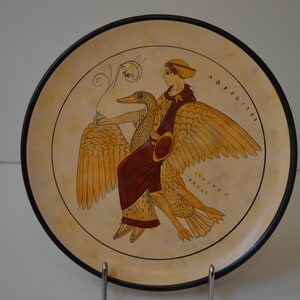 May include: A round ceramic plate with a black rim, featuring a painted image of a woman riding a goose. The woman is wearing a red robe and a white hat. The goose has yellow feathers and a long neck. The plate has the words "APHRODITE" and "ALEXANDROS NAVIS" written in Greek around the image.