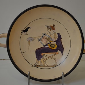 May include: A ceramic plate with a black rim and a cream-colored center. The plate features a hand-painted design of a woman playing a lyre, a black bird, and a floral motif. The plate has two handles.