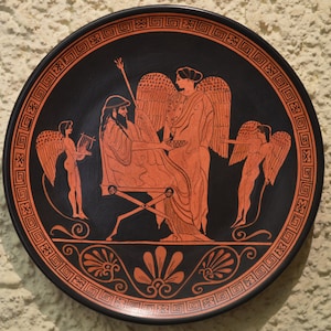 May include: A black and red ceramic plate with a Greek mythological scene. The plate features a seated figure with a lyre, a standing figure with wings, and two winged figures. The plate is decorated with a black and red geometric border.