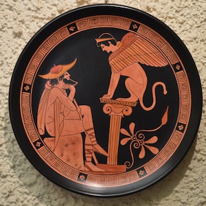 May include: A black ceramic plate with a red and white design depicting a winged creature and a seated figure. The plate has a black border with a repeating geometric pattern.