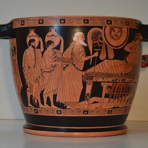 May include: A black and orange ceramic vase with two handles. The vase features a detailed illustration of ancient Greek figures in orange against a black background. The figures are engaged in various activities, including carrying objects and interacting with each other.