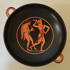 Può includere: Un piatto in ceramica nero con due figure dipinte in rosso e arancione. Le figure ballano e suonano un flauto. Il piatto ha due manici.