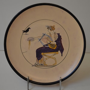 May include: A round ceramic plate with a black rim features a hand-painted design of a woman playing a lyre. The woman is wearing a purple robe and a crown of leaves. A black bird is perched on a branch to the left of the woman.