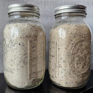 May include: Two clear glass mason jars filled with a light-colored granular substance. The jars have silver lids and are labeled with the word "Ball" and measurements on the side. The jars sit on a black surface.