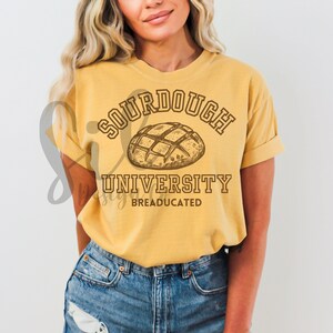 Può includere: Una maglietta gialla con il testo "Sourdough University" e un disegno di una pagnotta di pane a lievitazione naturale. Il testo "BREADUCATED" è sotto il nome dell'università.