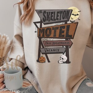 May include: A beige sweatshirt featuring a vintage motel sign graphic that reads "The Skeleton Motel" with the words "Free Nightmares" and "Vacancy" on the sign. A pumpkin and a skull are depicted on the sign.