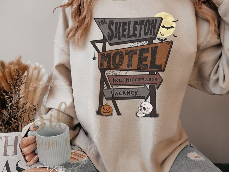 May include: A beige sweatshirt featuring a vintage motel sign graphic that reads "The Skeleton Motel" with the words "Free Nightmares" and "Vacancy" on the sign. A pumpkin and a skull are depicted on the sign.