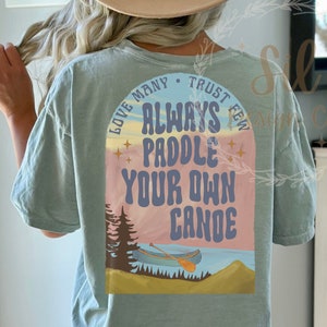 May include: A light blue t-shirt with a graphic print of a sunset scene with a canoe and the text "Love Many, Trust Few, Always Paddle Your Own Canoe".