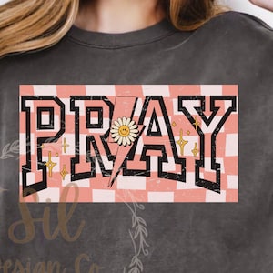 May include: A grey sweatshirt with a pink and white chequered background and the word "PRAY" in black block letters. A yellow daisy with a smiley face is in the centre of the word "PRAY".