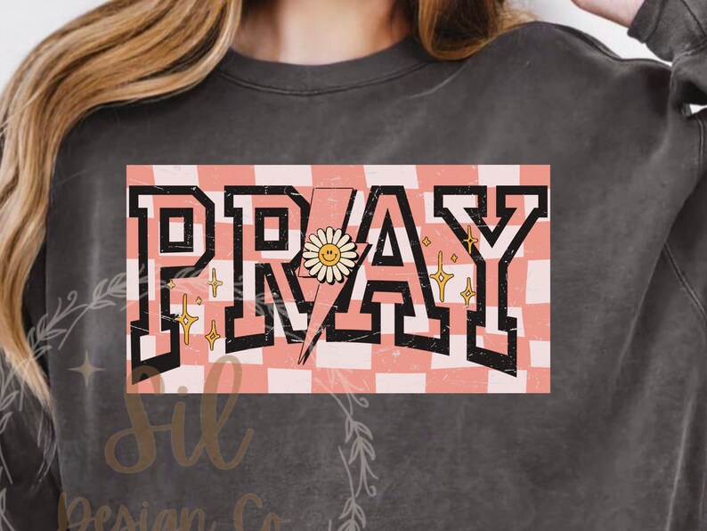 May include: A grey sweatshirt with a pink and white chequered background and the word "PRAY" in black block letters. A yellow daisy with a smiley face is in the centre of the word "PRAY".
