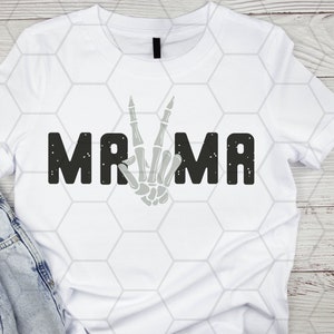 Puede incluir: Camiseta blanca con un gr&aacute;fico negro que dice "MAMA" con una mano de esqueleto haciendo un signo de paz en el medio de la palabra.