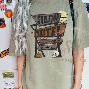 May include: A green t-shirt featuring a vintage-style graphic of a motel sign that reads "The Skeleton Motel". The sign has a pumpkin and skull underneath it. The text on the sign reads "Free Nightmares" and "Vacancy".
