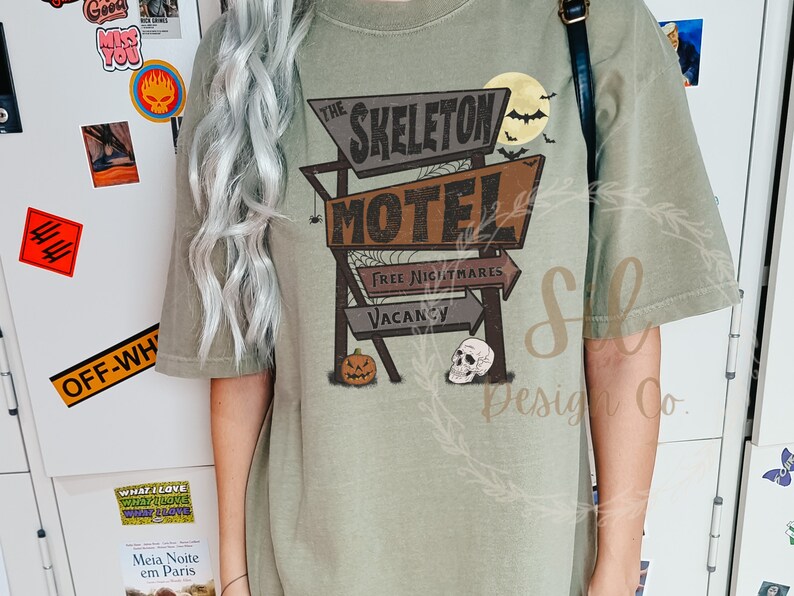 May include: A green t-shirt featuring a vintage-style graphic of a motel sign that reads "The Skeleton Motel". The sign has a pumpkin and skull underneath it. The text on the sign reads "Free Nightmares" and "Vacancy".
