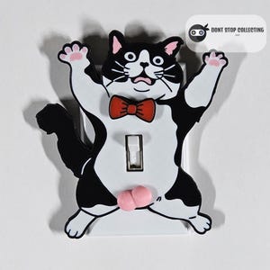 May include: A black and white cat wearing a red bow tie is depicted on a white light switch cover. The cat has a surprised expression and its paws are raised in the air. The words "Don't Stop Collecting Inc." are printed in the top right corner of the image.