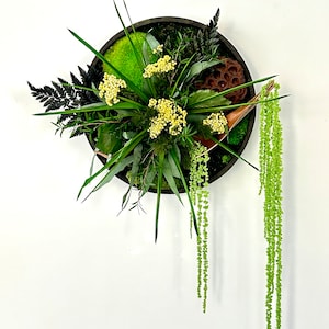 Può includere: Un'appendice murale circolare con una varietà di piante e fiori verdi, tra cui felci, muschio e un baccello di loto secco. L'appendice è incorniciata da un cerchio marrone scuro.
