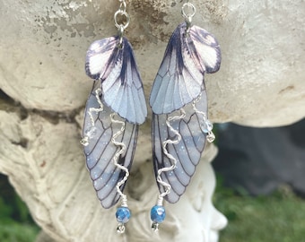 May include: A pair of dangle earrings featuring translucent, iridescent insect wing shapes in shades of pale blue and purple. Each earring includes a silver-toned wire accent in a zigzag pattern, a small clear bead, and a blue faceted bead at the base.