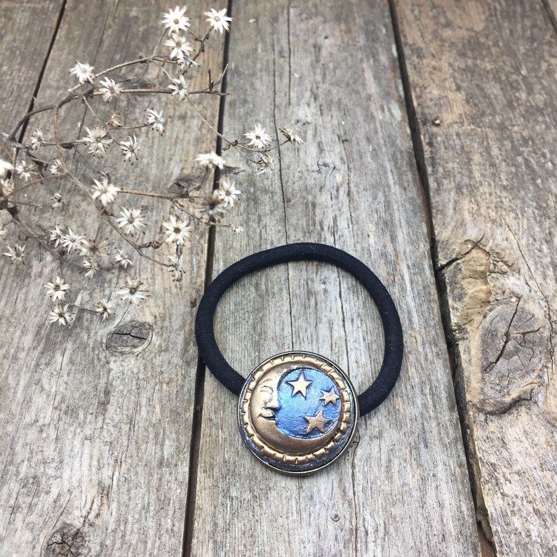 May include: A black elastic hair tie with a gold and blue crescent moon charm featuring stars. The charm has a textured, aged look.