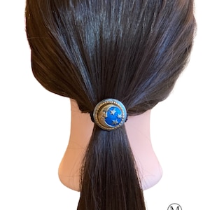 May include: A blue and gold crescent moon hair tie with stars. The hair tie is on a brown mannequin head with long brown hair.