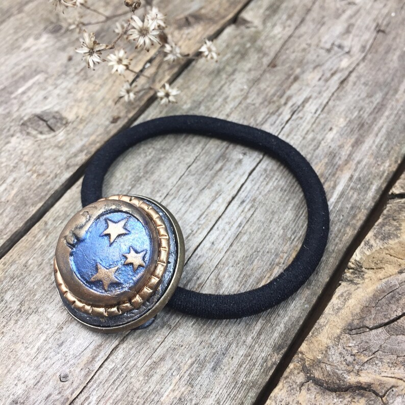 May include: A black hair tie with a gold and blue crescent moon charm with gold stars. The charm is attached to the hair tie.