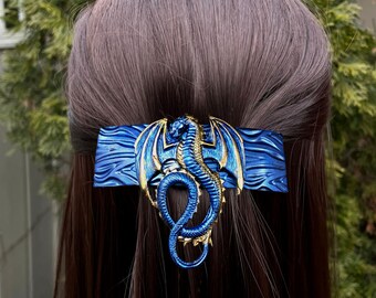 May include: Blue and gold dragon hair clip. The dragon is curled up with its wings spread out. The clip is made of metal and has a textured surface.