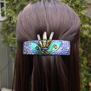 May include: A purple and green hair clip with a geometric pattern and iridescent crystal accents. The clip features a leaf design and a gold-colored accent.