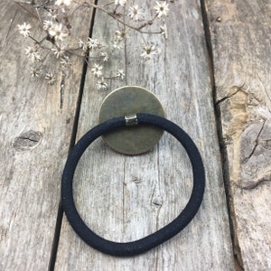 May include: A black hair tie with a round, bronze-colored metal accent. The accent has a small, silver-colored metal piece in the center.