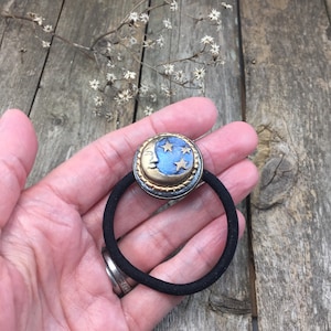 May include: A black elastic hair tie with a gold and blue crescent moon charm. The charm has a face and five stars.