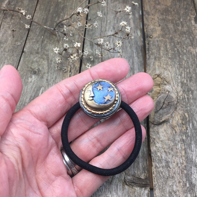 May include: A black elastic hair tie with a gold and blue crescent moon charm. The charm has a face and five stars.