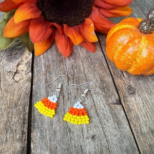 May include: A pair of dangle earrings featuring a triangular candy corn design crafted from small beads in horizontal stripes of white, orange, and yellow. Each earring is attached to a silver-toned metal hook for pierced ears.
