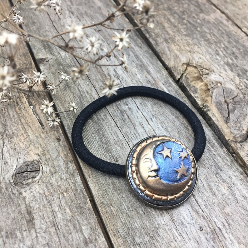 May include: A black elastic hair tie with a gold and blue crescent moon charm. The charm has a face and stars on it.