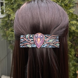 May include: A purple and copper owl with intricate details is the centerpiece of this handmade hair clip. The owl is surrounded by branches and leaves, creating a woodland scene. The clip is made of polymer clay and is perfect for adding a touch of whimsy to any hairstyle.