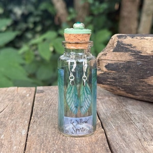 May include: A glass bottle with a cork stopper and a green gem. Inside, two silver earrings with green, wing-shaped pendants are displayed. The bottle has a label that reads "Willow Sprite".